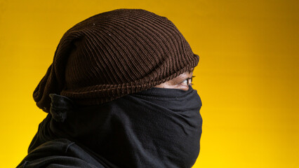 Striking profile portrait of a young Asian male wearing a black fabric mask and a brown knitted beanie set against a bright yellow background. The subject faces completely to the side, © M. Faisal Riza