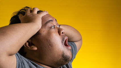 Dynamic profile portrait of a young Asian male expressing extreme frustration or panic against a vibrant yellow background. The distressed individual holds both hands tightly on the © M. Faisal Riza