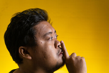 Expressive side profile portrait of a young Asian male making a recognizable silence gesture against a vibrant yellow background. The individual places a single index finger vertically © M. Faisal Riza