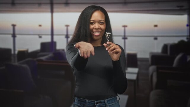 Woman holding eyelash curler offers palm and smiles in airport lounge with window view of sea at sunset; friendly welcome.