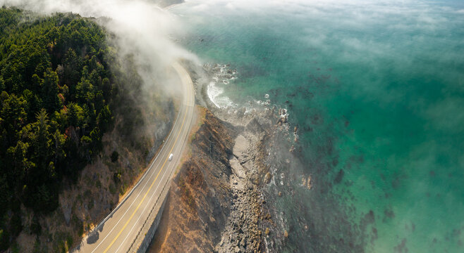 Aerial view of Highway 101 winding along the rugged Oregon coastline, with dense forests, rocky cliffs, and sea fog over the turquoise Pacific Ocean in Port Orford, Oregon, United States.