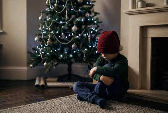 Sad young boy sitting on the floor near a decorated christmas tree