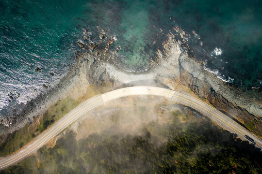 Aerial view of Highway 101 curving along the rugged coastline with rocky cliffs, crashing waves, and patches of fog over the forest in Port Orford, Oregon, United States.