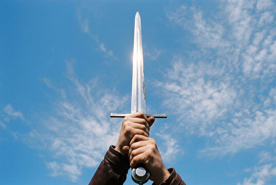 Two strong hands firmly holding a shiny medieval steel sword pointing upward toward a blue sky