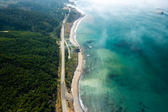Aerial view of Highway 101 winding along the rugged coastline with dense evergreen forests and turquoise ocean waters under a light coastal fog in Port Orford, Oregon, United States.