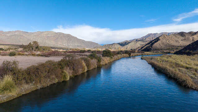 Aerial view of the San Juan River near the abandoned aluminum sulfate plant ruins in Calingasta, San Juan, Argentina, surrounded by mountains and shrubs under a clear blue sky.