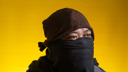 Striking profile portrait of a young Asian male wearing a black fabric mask and a brown knitted beanie, posing against a solid yellow background. The subject looks thoughtfully away © M. Faisal Riza
