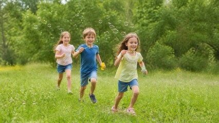 Fototapeta premium Group of happy children running on green lawn and blowing soap bubbles outdoors in summer park