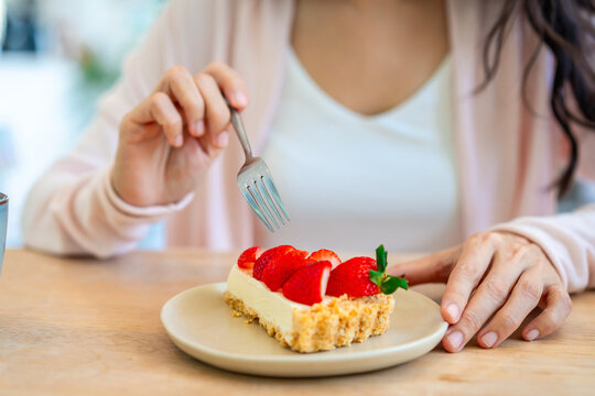 Cropped shot of a happy young woman holding a slice of strawberry cake and a fork, enjoying dessert in a cozy cafe