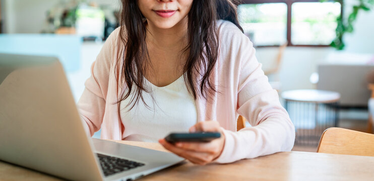Cropped shot of a thoughtful Asian woman working on a smartphone and laptop at a bright home desk