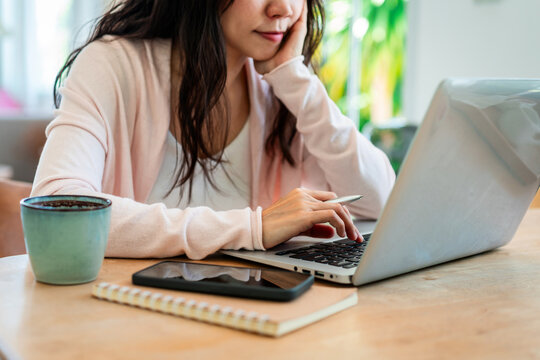 Cropped shot of a thoughtful Asian woman resting her chin on hand while working on laptop at a bright home desk