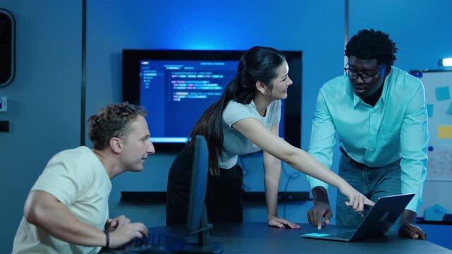 Diverse team of IT professionals collaborating on software development code late at night in a high-tech office. Software engineers pointing at laptop screen, discussing cybersecurity project.