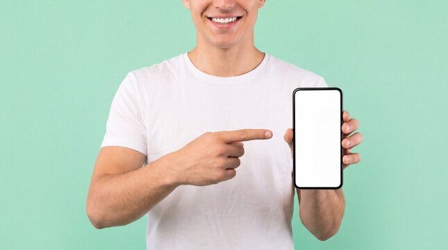 Man wearing white t-shirt holding black smartphone with blank white screen and pointing index finger to mobile on solid mint green background featuring phone mockup