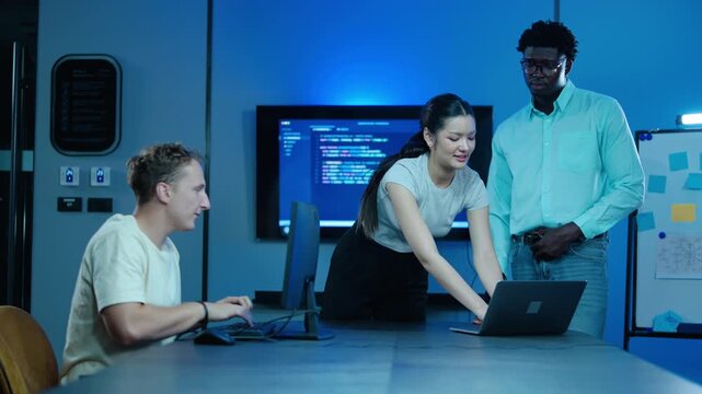Diverse team of IT professionals collaborating on a software project in a modern dark office workspace. Programmers analyzing code on laptops and desktop computers during a late night work session.
