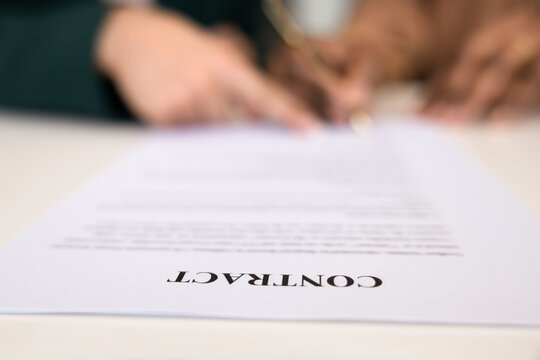 Close up view of printed agreement paper with word CONTRACT in focus, while two diverse blurred female hands in background signing, pointing at document. Agreement, partnership, official business deal