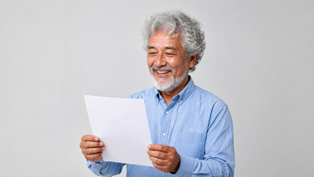 An elderly man with curly gray hair and a beard smiles while reading a white sheet of paper against a plain light background.