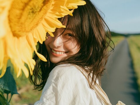 夏の青空が広がる田舎道で、大輪のひまわりの隣で眩しい笑顔を見せる女性