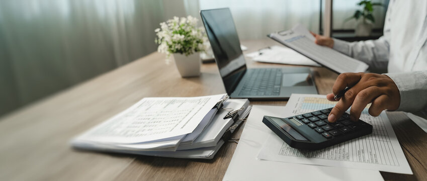 Person calculates figures on financial documents laptop while another reviews a clipboard, supporting account analysis, financial checking, business account management copy space