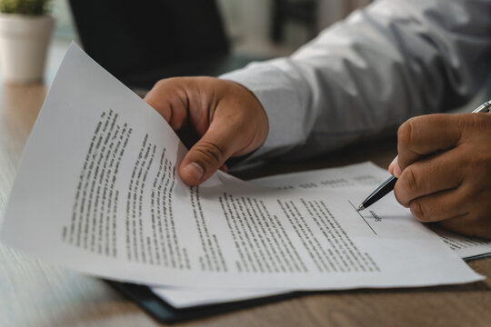 Close-up of hands reviewing printed documents and signing on a marked line with a pen at a desk, showing a contract approval process and business agreement workflow in a modern office.