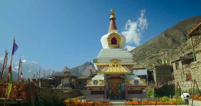 Annapurna Circuit, Manang Nepal,Traditional white and gold Buddhist chorten monument with prayer flags, near the Himalayan mountain peaks under a clear blue sky.