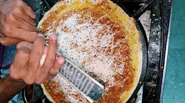 Close up of a street food vendor grating fresh cheese over a masala dosa on a hot tawa, preparing a crispy South Indian dish at a busy Indian food stall