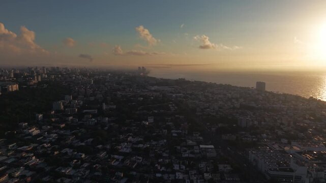 High-angle aerial drone 360-degree clockwise orbit shot over Santo Domingo city, showcasing the residential areas, Mirador Sur park, Anacaona skyline, and golden sunrise over the Caribbean sea