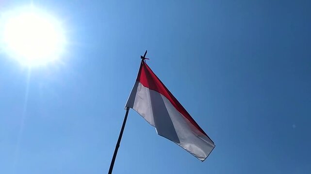 The red and white Indonesian national flag waves ahead of Independence Day, set against a backdrop of sunlight and a clear daytime sky.