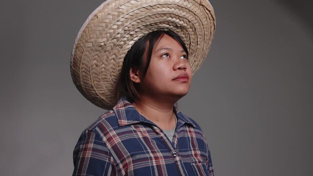 Cheerful young Asian female farmer in a straw hat and plaid shirt laughing and looking up with joy.