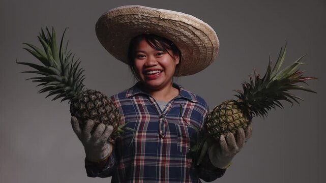 Happy young Asian female farmer wearing a straw hat and plaid shirt, holding fresh pineapples in both hands.