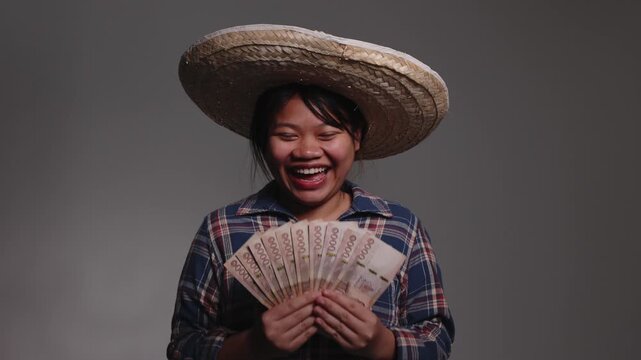 Happy young Asian female farmer wearing a straw hat and plaid shirt, holding a fan of Thai baht banknotes.