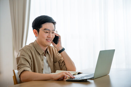 Happy young Asian man working on laptop in living room. Freelance worker male sitting at desk writing notes while watching webinar, studying online
