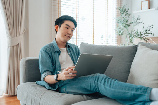 Young Asian man relaxing on comfortable couch, using laptop at home, happy male chatting with friends in social network, spending weekend, watching movie, shopping online