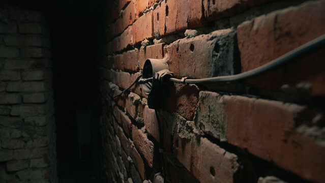 Dimly lit basement corner featuring aged red brick wall with exposed electrical conduit metal junction box. Warm spotlight creates dramatic shadows highlights texture