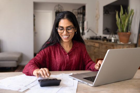 Smiling Indian woman with glasses using calculator and online banking on laptop while reviewing bills and receipts, managing personal or family expenses, planning finances and budgeting at modern home