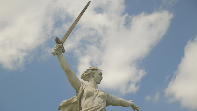 Monumental stone statue of female figure raising sword high, dynamic pose against blue sky with scattered clouds. Strong backlighting highlights form. Evokes courage defiance