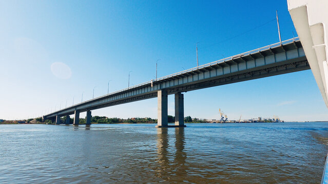 Low angle view of large concrete bridge over calm river under bright blue sky. Industrial cranes visible on far bank. Strong diagonal composition, natural daylight, cool tones