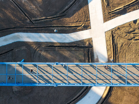 Aerial view of a blue pedestrian bridge with people walking across it, set against a backdrop of dirt paths and construction work in Belgrade, Serbia.