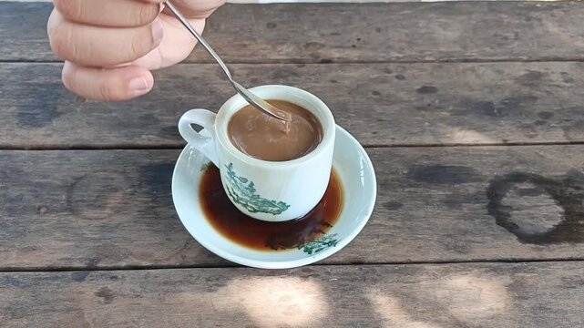 Video clip of a hand stirring traditional milk coffee in a ceramic cup on a rustic wooden table.