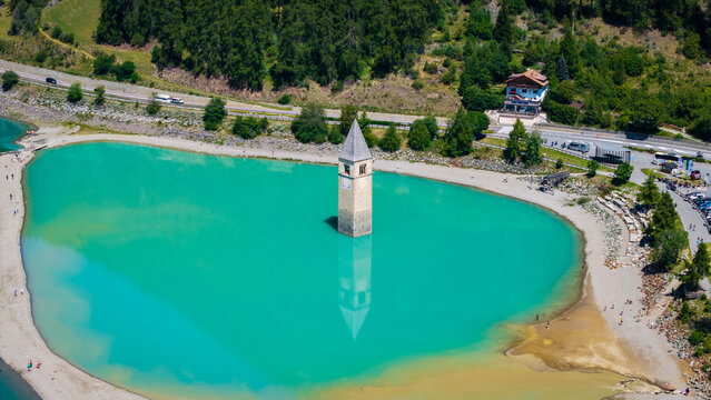 Aerial view of the submerged bell tower of Curon rising from the turquoise waters of Lake Reschen, surrounded by green mountains in Reschen am See, Trentino-Alto Adige, Italy.