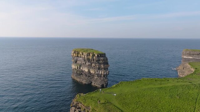 Dun Briste sea pillar sits just off a flat coastal headland as the drone moves slowly along the Mayo shoreline in bright daytime.