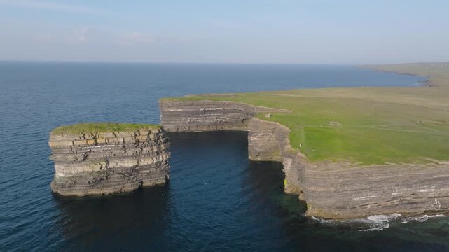 Layered rock column Dun Briste stands off a flat grassy cliff top above calm Atlantic waters in bright daytime light.