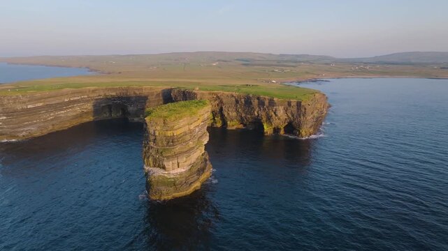 Camera glides past Dun Briste sea stack and arched coastal wall with rural fields in warm evening light.