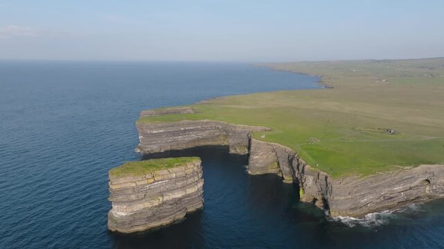 Layered rock column Dun Briste stands off a flat green headland above calm Atlantic water, viewed from a high circling drone.