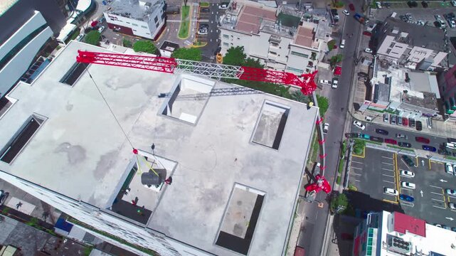 High-angle aerial drone top-down shot pulling back over a red tower crane lifting construction materials into a skyscraper rooftop opening in Naco, Santo Domingo