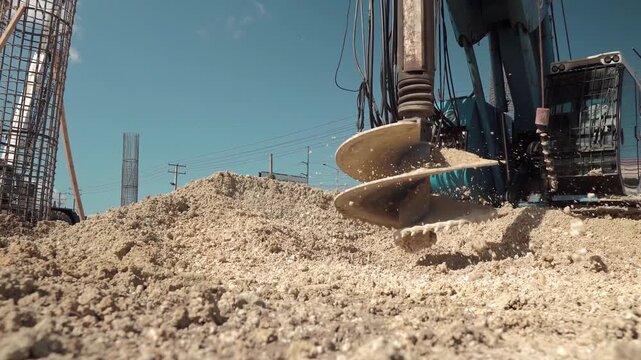 Low-angle ground-level static shot of a hydraulic piling rig's auger drill spinning rapidly to discharge soil onto a dirt pile at a civil engineering construction site
