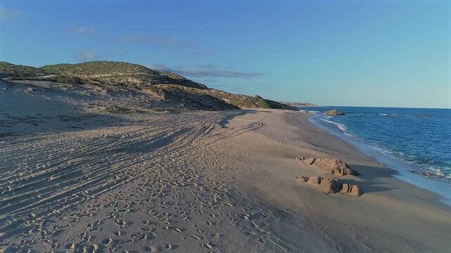 Drone continues flying low along the beach at Playa El Rinconcito, passing a small but obvious rock stuck in the sand and faint tire tracks along the shore before moving over the ocean as a wave rolls