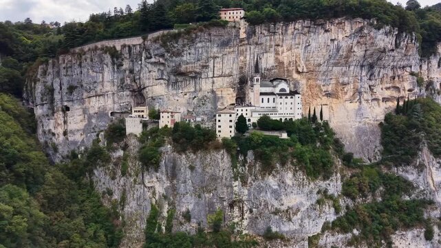 Wide drone view of Santuario Madonna della Corona built into a towering limestone cliff surrounded by lush forest in northern Italy