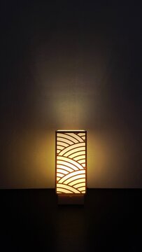 A tall, slender lamp glows warmly in an otherwise dark room. Its rectangular shade is cut with a scalloped, wave-like pattern of overlapping arcs reminiscent of traditional Japanese seigaiha