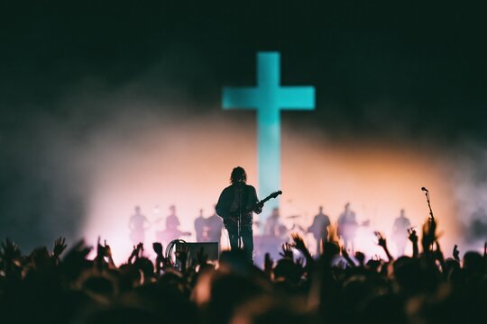 Luminous blue cross behind a silhouetted guitarist and band on a glowing stage, featuring a cheering crowd for worship and church media