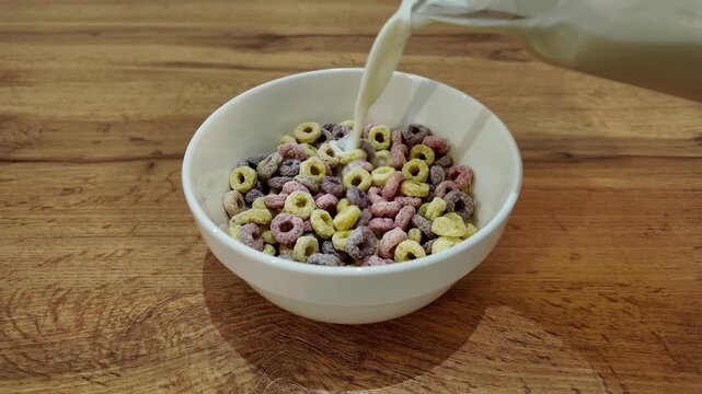Delicious and healthy breakfast preparation. Fresh milk pouring from a pitcher into a white bowl filled with colorful cereal rings on a wooden table
